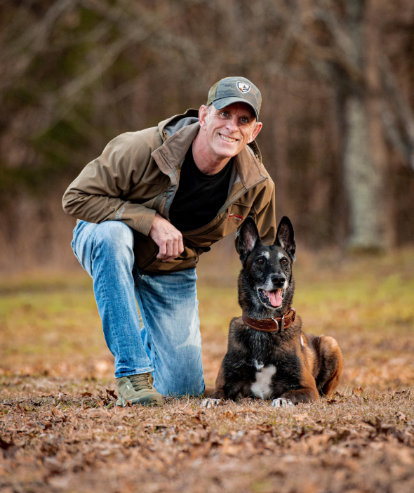 Phil Goldstine, retired U.S. Army Ranger and First Sergeant, kneeling beside a Belgian Malinois on the Supreme Canine estate grounds
